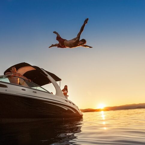 Friends enjoying summertime by boating on the lake. They watch as a young man dives spread eagle into the water at sunset.